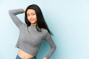 Genç Çinli bir kadın mavi bir arka planda izole edilmiş kafasının arkasına dokunuyor, düşünüyor ve bir seçim yapıyor..