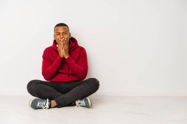 Young latin man sitting on the floor isolated laughing about something, covering mouth with hands.