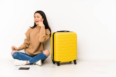 Genç Çinli gezgin kadın, izole edilmiş bir bavulla yerde oturuyor. Parmaklarıyla bir cep telefonu hareketi gösteriyor..