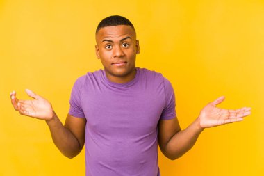 Sarı arka planda izole edilmiş genç latin adam sorgulama sırasında omuz silkiyor ve kuşkulanıyor..