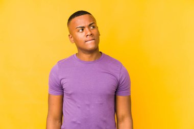 Sarı arka planda izole edilmiş genç latin adam kafası karışmış, şüpheli ve güvensiz hissediyor..