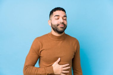 Mavi arka planda genç bir latin adam göbeğe dokunur, nazikçe gülümser, yer ve tatmin olur..