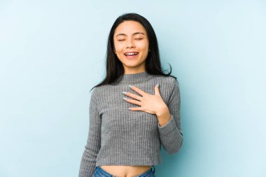 Mavi arka planda izole edilmiş genç Çinli kadın elini göğsünde tutarak kahkahalar atıyor..