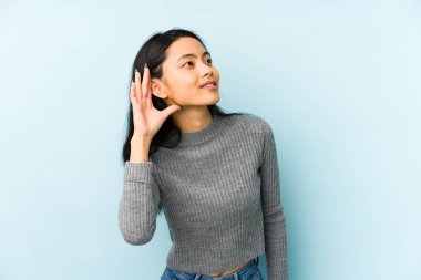 Genç Çinli bir kadın mavi arka planda izole edilmiş bir şekilde bir dedikoduyu dinlemeye çalışıyor..