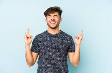 Mavi arka planda izole edilmiş genç bir Arap, parmaklarıyla kaya hareketi gösteriyor.