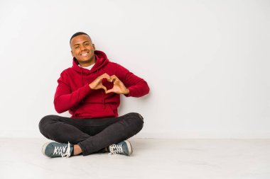 Yerde tek başına oturan genç Latin adam gülümsüyor ve elleriyle kalp şeklini gösteriyor..