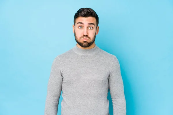 Young caucasian man against a blue background isolated blows cheeks, has tired expression. Facial expression concept.