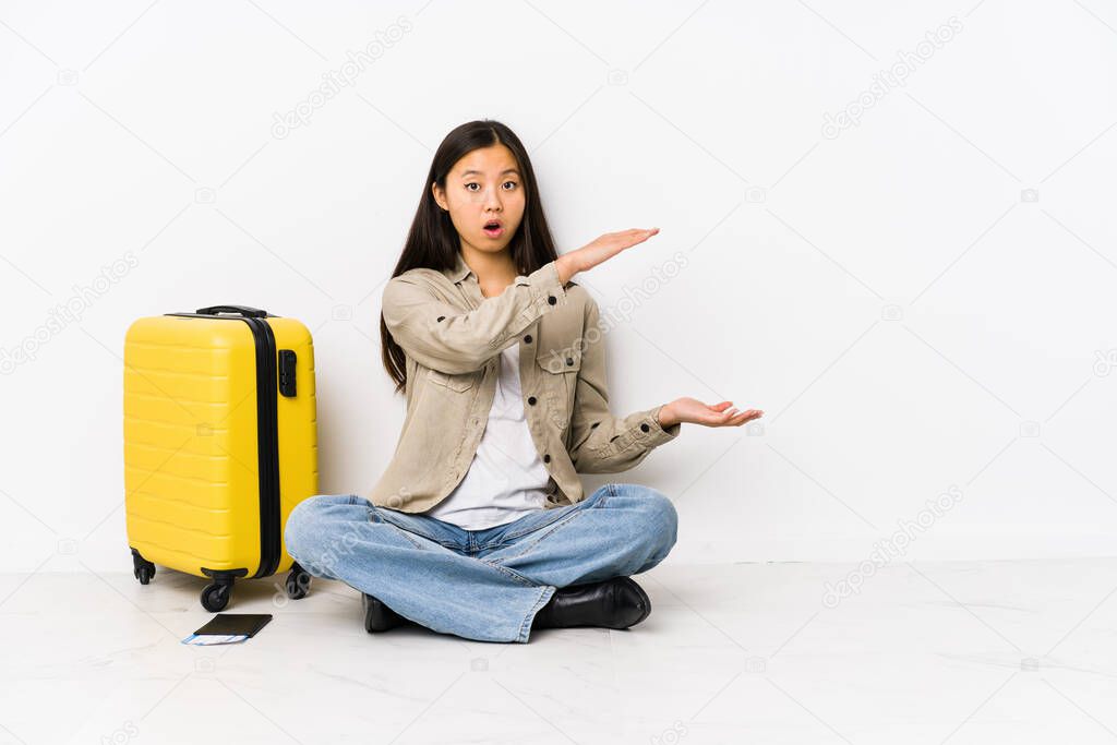 Young chinese traveler woman sitting holding a boarding passes shocked and amazed holding a copy space between hands.