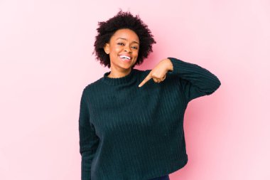 Orta yaşlı Afro-Amerikalı bir kadın pembe arka planda tek başına bir gömlek fotokopi alanını işaret eden gururlu ve kendinden emin birine karşı.