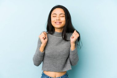 Mavi arka planda izole edilmiş genç Çinli kadın mutlu ve başarılı hissediyor. Zafer konsepti.