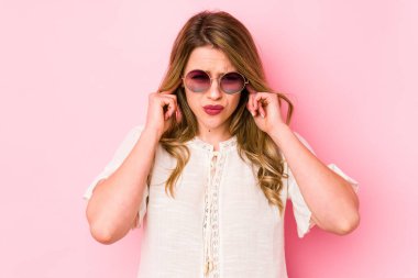 Pembe arka planda gözlüklü genç beyaz kadın kulaklarını parmaklarıyla kapatıyor, gürültülü bir ortam yüzünden stresli ve çaresiz..