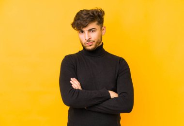Sarı arka planda izole edilmiş, hoşnutsuzca kaşlarını çatmış, kollarını kavuşturmuş genç bir Arap..