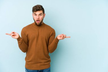 Mavi arka planda izole edilmiş genç beyaz adam farklı kopya alanlarını gösteriyor, birini seçiyor, parmaklarıyla gösteriyor.