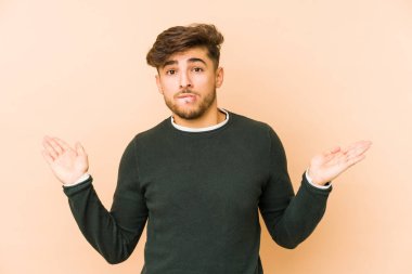 Bej bir arka planda izole edilmiş genç Arap bir adam. Kopya alanını tutmak için kafası karışık ve omuz silkiyor..