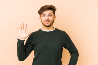 Genç Arap bir adam bej bir arka planda izole edilmiş. Gülümsüyor beş numarayı parmaklarıyla gösteriyor..