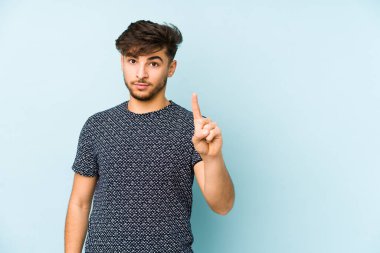 Genç Arap bir adam mavi arka planda izole edildi. Parmağıyla bir numarayı gösteriyor..