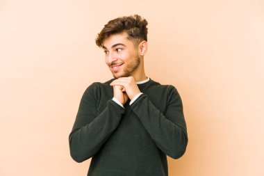 Bej bir arka planda izole genç Arap adam ellerini çenesinin altında tutuyor, mutlu bir şekilde yana bakıyor..