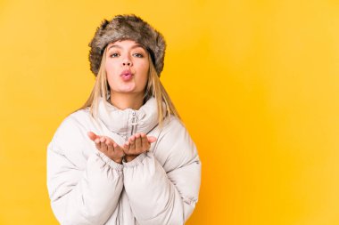 Kışlık kıyafetler giyen genç sarışın kadın sarı arka planda izole edilmiş dudakları kıvrılmış ve hava öpücüğü yollamak için avuç içleri tutan genç sarışın kadın..