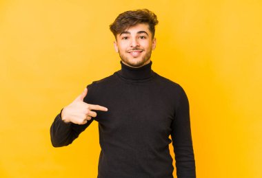 Sarı arka planda izole edilmiş genç bir Arap adam gömlek fotokopi alanını elle gösteriyor, gururlu ve kendinden emin.