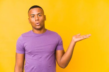 Sarı arka planda izole edilmiş genç latin adam, avuç içi fotokopi alanını tutmaktan etkilenmiş..