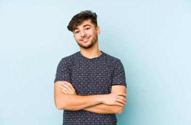 Mavi bir arka planda izole edilmiş genç bir Arap adam kendini güvende hissediyor, kararlılıkla kollarını kavuşturuyor..
