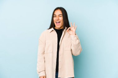 Mavi bir arka planda izole edilmiş genç bir kadın bir göz kırpıyor ve elleriyle iyi bir jest yapıyor..