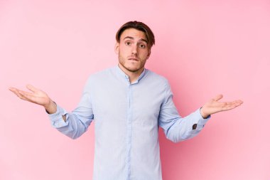 Pembe arka planda poz veren beyaz bir genç. Kafası karışmış ve omuz silkiyor..