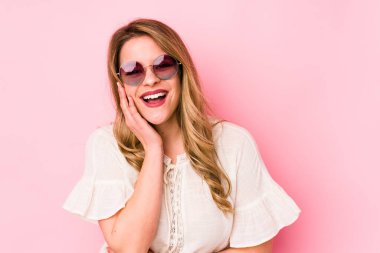 Pembe arka planda gözlüklü genç beyaz kadın mutlu bir şekilde gülüyor ve ellerini karnının üstünde tutarak eğleniyor..