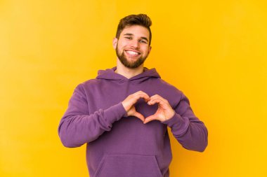 Sarı arka planda izole edilmiş genç beyaz adam gülümsüyor ve elleriyle kalp şeklini gösteriyor..