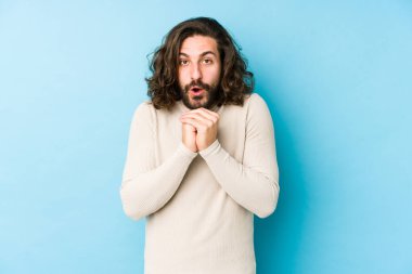 Uzun saçlı genç adam mavi bir arka planda izole edilmiş şans için dua ediyor, şaşırmış ve önüne bakıyor..