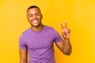 Sarı arka planda izole edilmiş genç latin adam zafer işareti gösteriyor ve gülümsüyor..