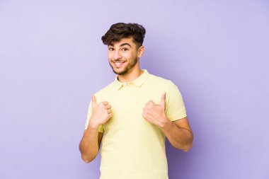 Mor bir arka planda izole edilmiş genç bir Arap adam iki parmağını da kaldırıyor, gülümsüyor ve kendinden emin..
