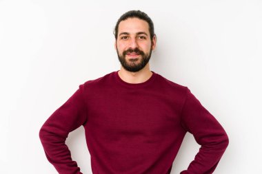 Genç, uzun saçlı, beyaz bir arka planda izole edilmiş, kendine güvenen, ellerini kalçasında tutan bir adam..