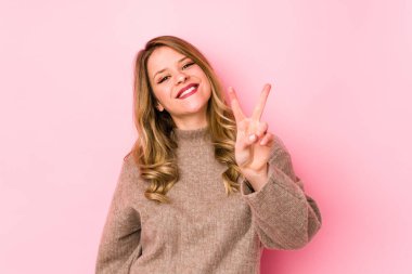 Pembe arka planda izole edilmiş genç beyaz kadın iki numarayı parmaklarıyla gösteriyor..