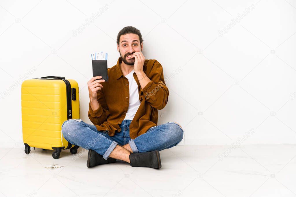 Young caucasian man sitting holding a passport and a suitcase isolated biting fingernails, nervous and very anxious.