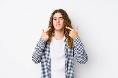 Uzun saçlı genç adam, yalnız gülümsüyor, parmaklarını ağzına doğrultuyor..