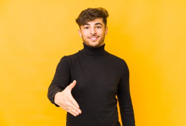 Sarı arka planda izole edilmiş genç bir Arap adam selamlama jesti olarak kameraya elini uzatıyor..