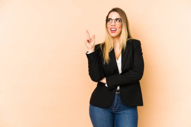 Orta yaşlı iş kadını, bej arka planda izole edilmiş, mutlu bir şekilde işaret parmağıyla işaret ederek gülümsüyor..