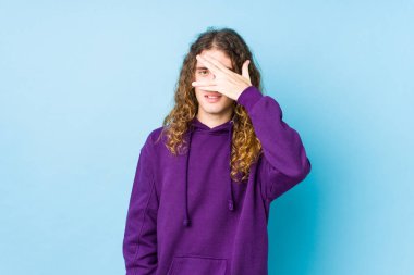 Uzun saçlı adam kameraya parmaklarıyla göz kırpıyor, utangaç bir şekilde yüzünü kapatıyor..