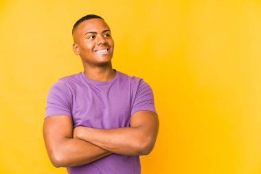 Sarı arka planda izole edilmiş genç Latin adam kollarını kavuşturarak gülümsüyor..