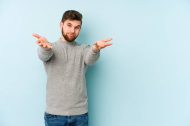 Mavi arka planda izole genç beyaz adam kollarıyla ölçek yapar, mutlu ve kendinden emin hisseder..