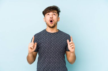 Mavi arka planda izole edilmiş genç bir Arap adam ağzı açık bir şekilde üst tarafı işaret ediyor..