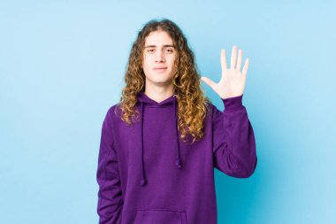 Uzun saçlı adam yalnız poz veriyor. Gülümsüyor. Beş numarayı parmaklarıyla gösteriyor..