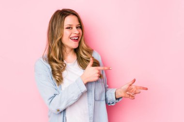 Pembe arka planda izole edilmiş genç beyaz kadın işaret parmaklarıyla işaret ederek heyecanlanıyor..
