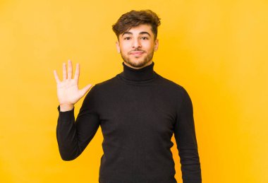 Sarı arka planda izole edilmiş genç bir Arap adam gülümseyerek beş numarayı parmaklarıyla gösteriyor..