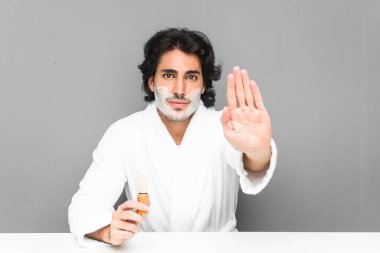 Genç bir adam sakalını tıraş ediyor. Elini uzatmış, dur işareti gösteriyor ve seni engelliyor..