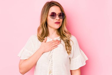 Pembe arka planda gözlüklü genç beyaz kadın yemin ediyor, elini göğsüne koyuyor..