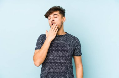 Mavi arka planda izole edilmiş genç bir Arap adam esniyor. Ağzı eliyle kapatan yorgun bir jest gösteriyor..