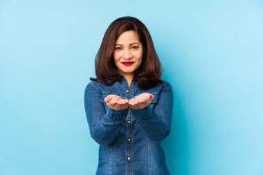 Mavi arka planda izole edilmiş orta yaşlı Latin bir kadın elinde avuç içi bir şeyle kameraya sunuyor..