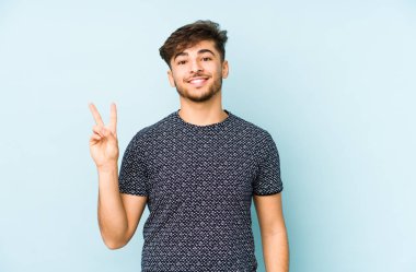 Genç Arap adam mavi bir arka planda izole edilmiş neşeli ve kaygısız parmakları olan bir barış sembolü gösteriyor..